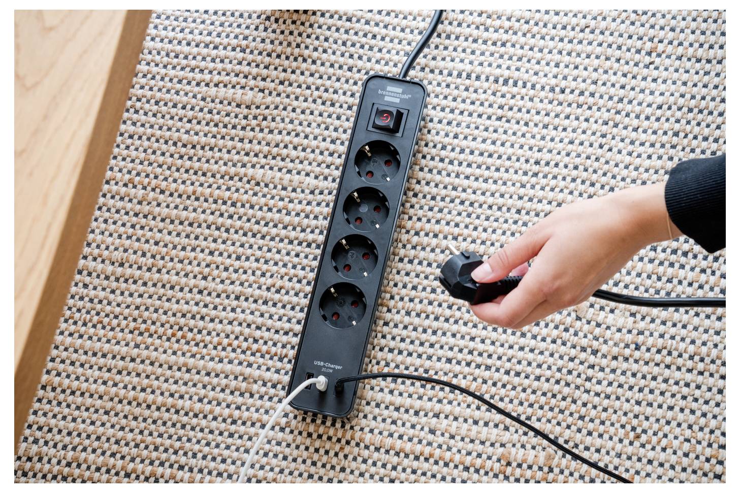 Egy személy keze egy kábelét csatlakoztatja egy fekete elosztóba négy csatlakozóaljzattal, amely egy textúrált szőnyegre van helyezve.
