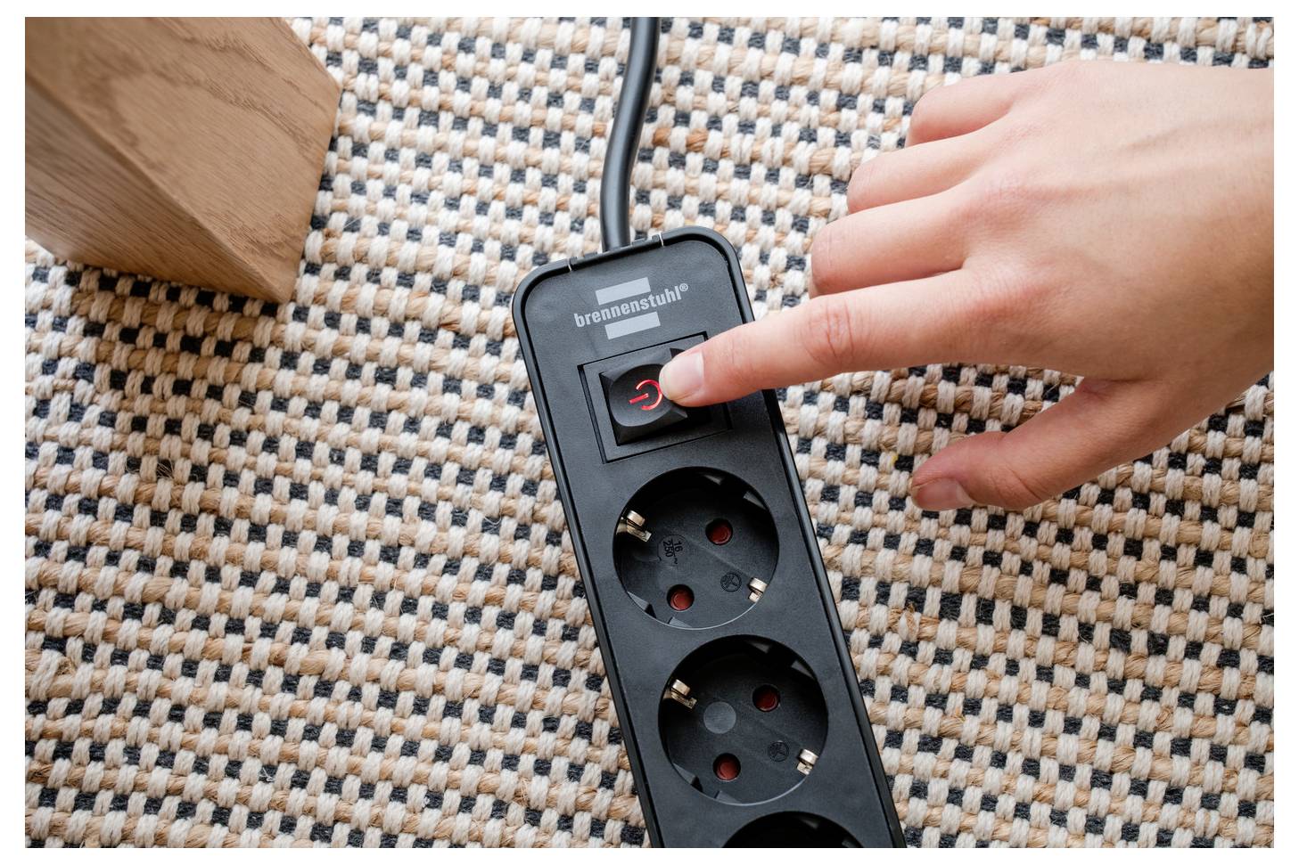 Egy kéz megnyomja a "power" gombot egy fekete elosztón, amely egy szövött szőnyegen helyezkedik el, egy fa bútor láb mellett.