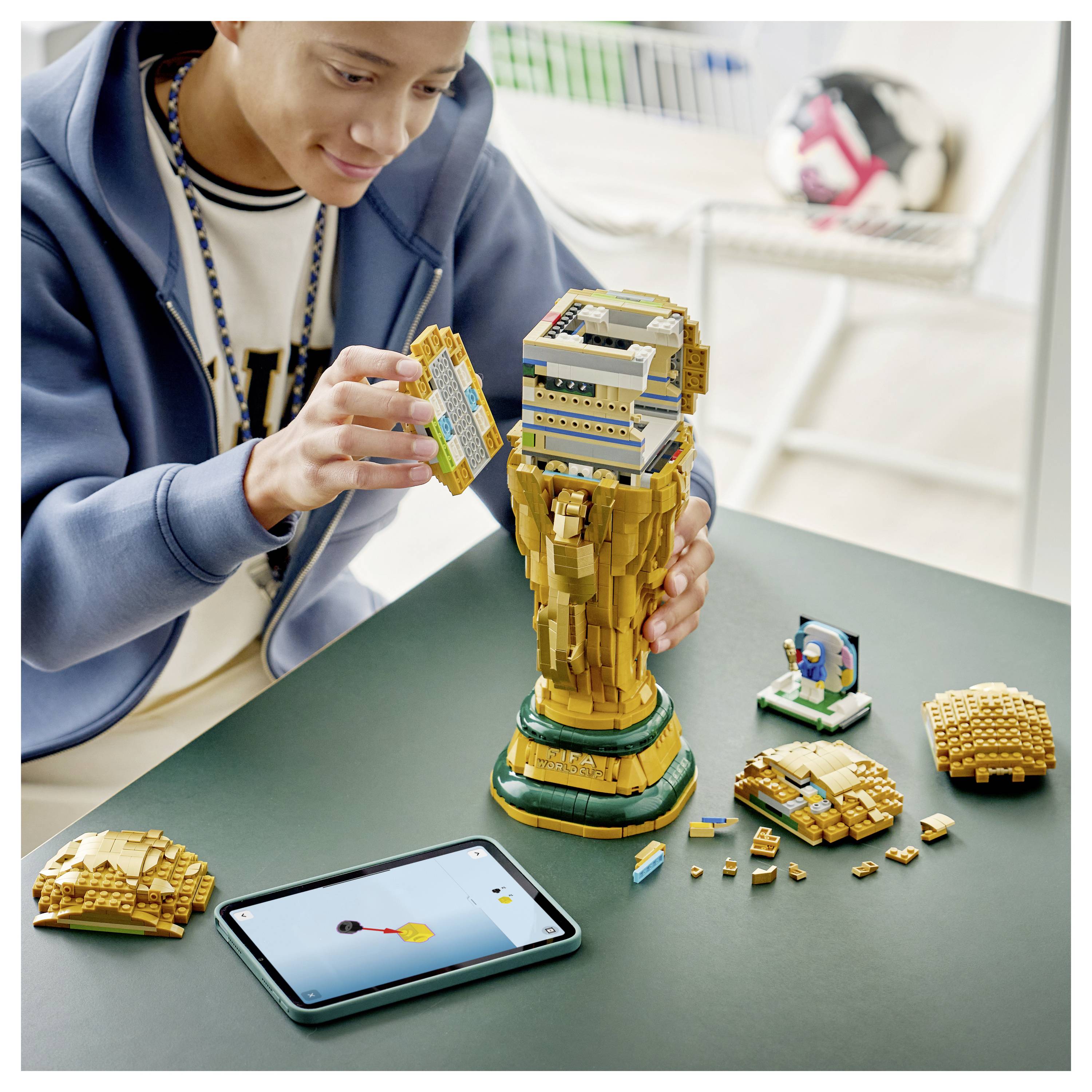 Egy személyt ábrázoló kép egy magas LEGO trófea modell összeszereléséről egy asztalon, egy tableten megjelenítendő animáció mellett. Szétszóródott LEGO alkatrészek vannak a közelben.