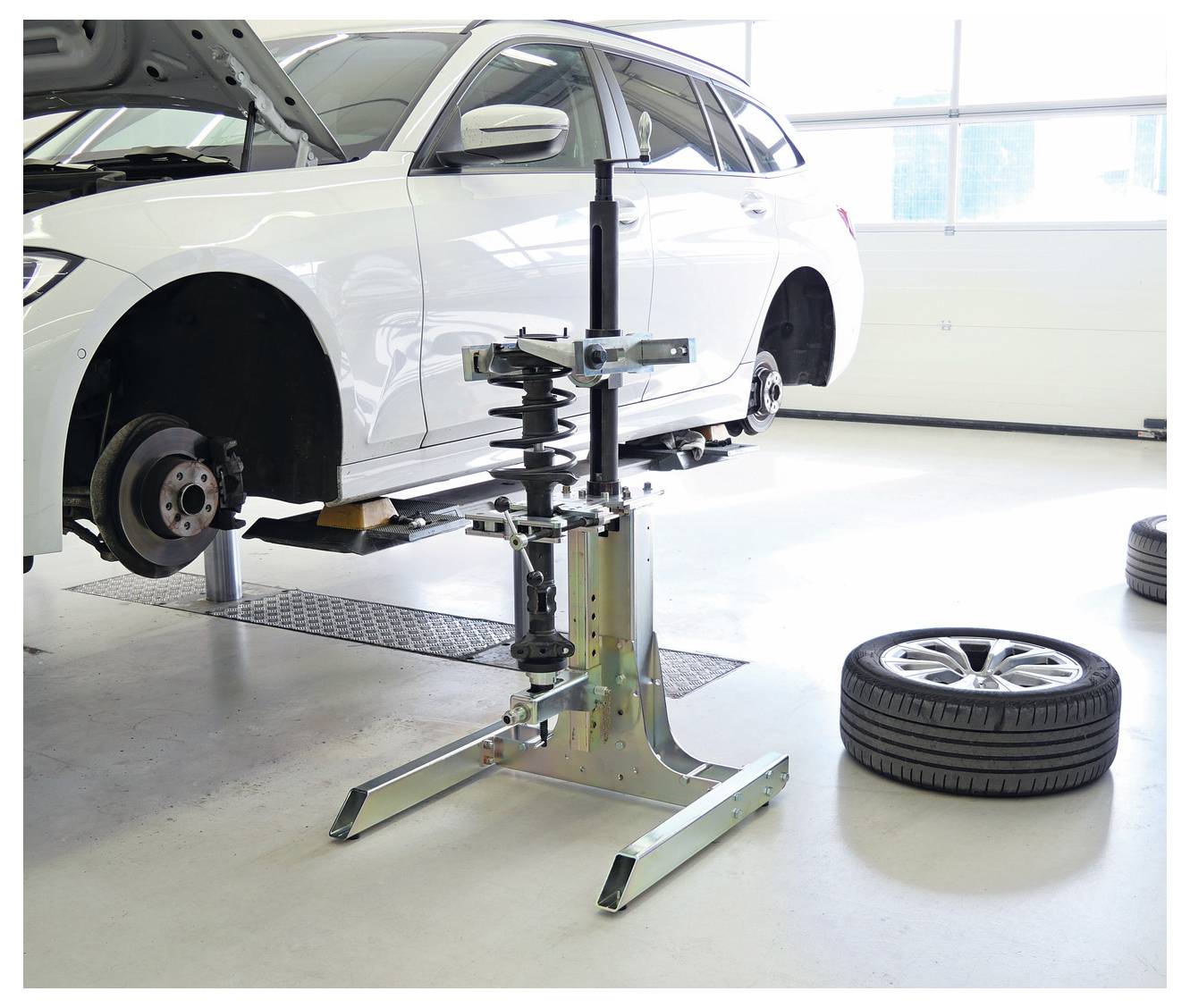Egy fehér autó egy benzinkúton van felmagasztva. Az első kereke le van szerelve. Közelében egy abroncscsere gép áll készen, egy pótkerék pedig a padlón fekszi.
