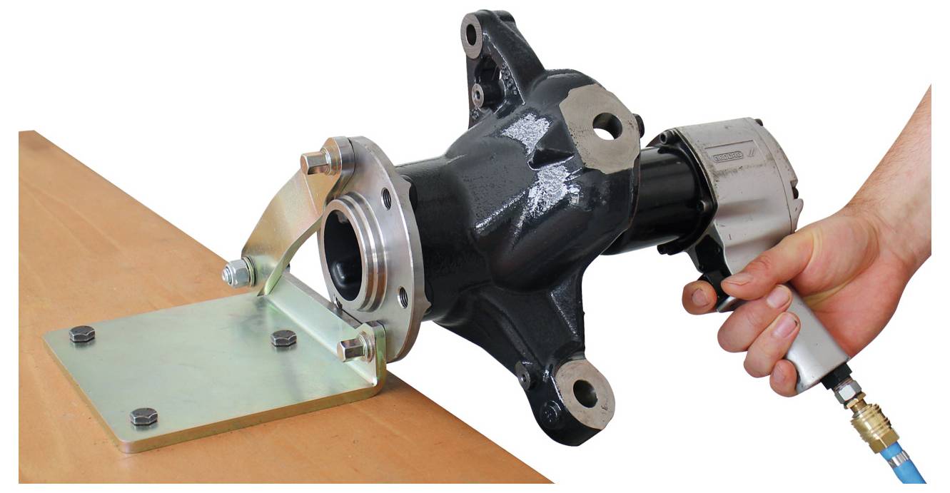 Egy kéz egy szerszámmal egy nagy fekete mechanikai alkatrészt rögzít egy fém lemezre, amely egy fa felületre van csatlakoztatva.
