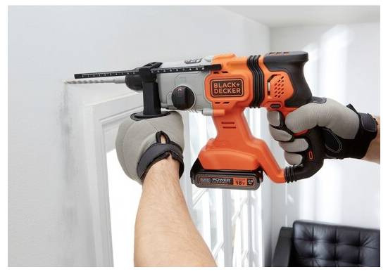 Egy személy narancsszín és fekete vezeték nélküli elektromos fúrót használ egy fal ablak közeli részébe lyuk fúrásához. A kezek védőkesztyűt viselnek.