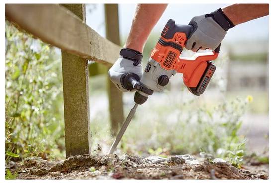 Egy kesztyűs személy egy narancs és fekete elektromos fúrógépet használ egy fa oszlop talajban való rögzítésére, füvet és napfényt körülvevően.