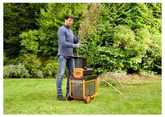 Egy személy kerti aprítógépet használ ágak felaprításához, fűvel borított terepen áll, a háttérben sűrű zöld lombozat látható.