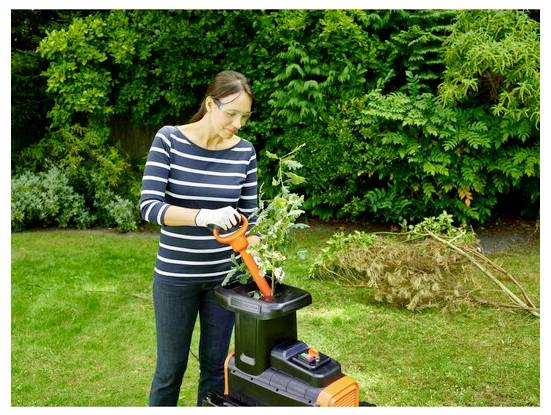 Egy csíkos ingben lévő nő elektromos kerti aprítót használ ágakon egy kertben, amely buja zöld fákkal és füvekkel van körülvéve.
