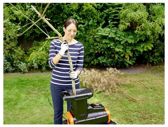 Egy biztonsági szemüveget és kesztyűt viselő személy ágakat táplál egy kertészeti aprítógépbe, a háttérben zöldséggel és száraz ágakkal.