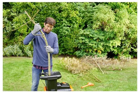 Kesztyűt viselő férfi ágakat táplál egy kerti aprítógépbe, amely zöldséggel és nyírott ágakkal van körülvéve egy kertben.