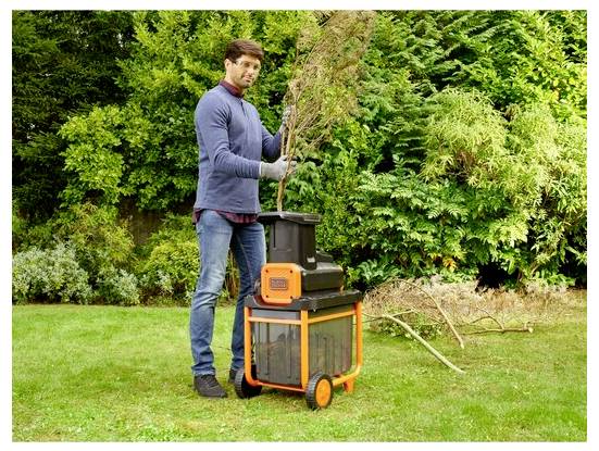 Egy személy elektromos ágaprítót használ az ágak feldolgozásához egy kertben, amely üde zöld lombozattal van körülvéve.