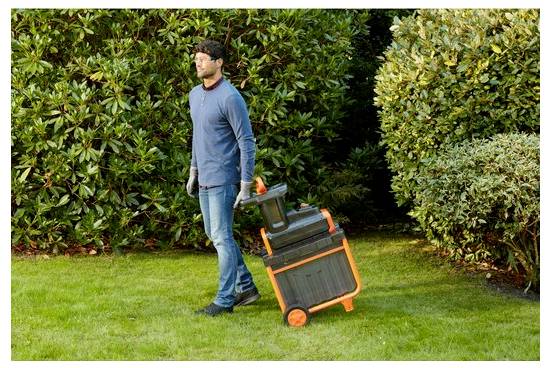 Egy könnyedebb ruházatban és kesztyűben öltözött személy egy lombszívót húz végig egy füves terülten, amelyet sűrű cserjék vesznek körül.