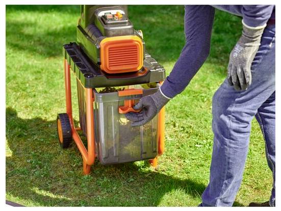 Egy kesztyűt viselő személy egy narancssárga és fekete lombshreddelőt működtet füvön, ami a kerti munkára és törmelékkezelésre utal.