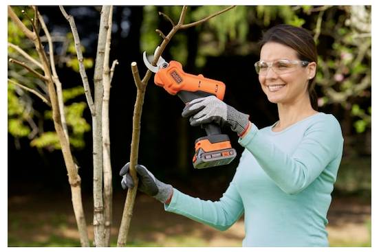 Egy szemüveget és kesztyűt viselő személy egy narancssárga akkumulátoros fűrészt használ egy kis fa ágainak nyírásához egy kertészeti környezetben.
