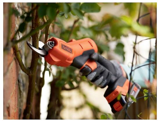 Személy elektromos metszőszerszám használatával ágakat vágva egy kertben, védőkesztyűt viselve.