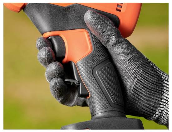 Egy kesztyűs kéz egy narancssárga és fekete elektromos szerszám, valószínűleg egy fúrógép fogantyúját fogja, homályos füves háttérrel a szabadban.