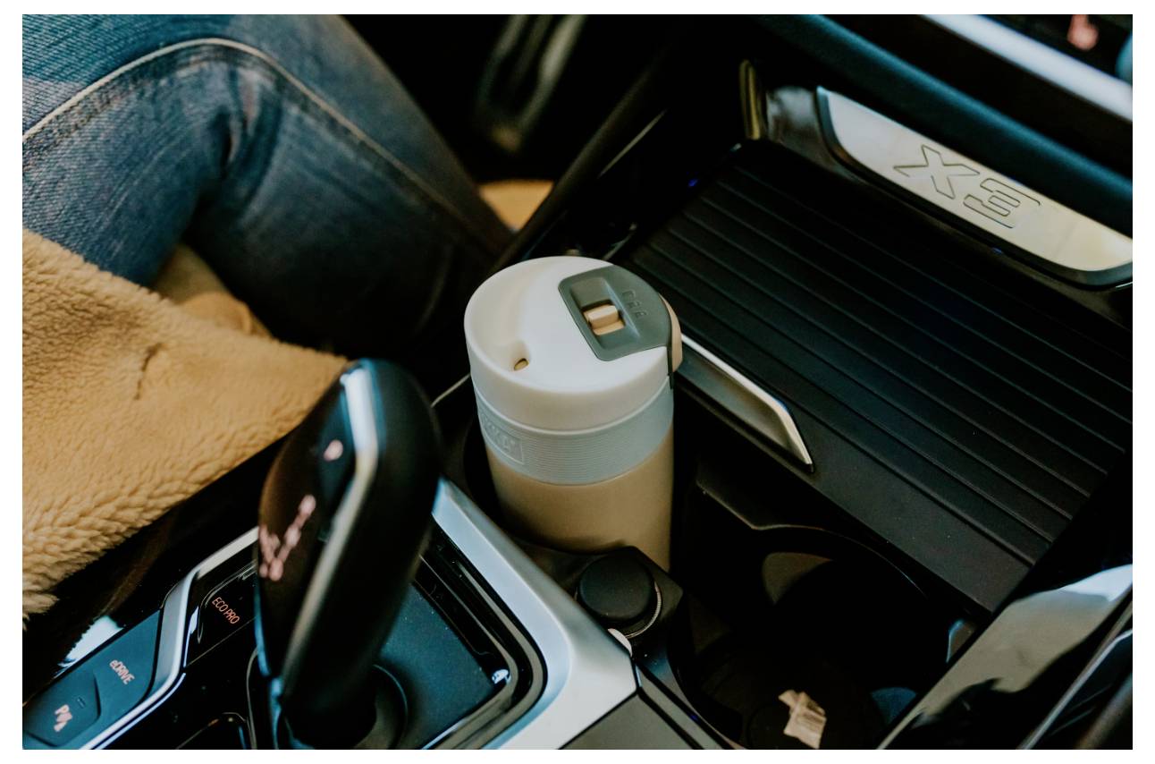 Egy utazási pohár az autó pohártartójában a váltó mellett, mellette egy farmer- és bézs szövetben ülő személy.