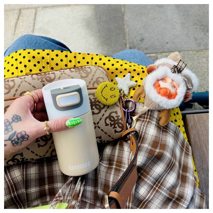 Egy tetoválással díszített kezű személy egy bézs Kambukka palackot és egy mintás táskát tart, amely különféle kulcstartókkal, köztük egy textil gombával van díszítve.