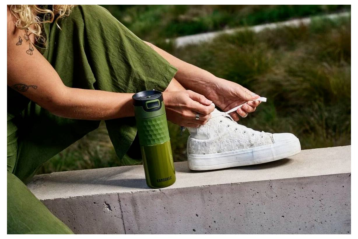 Egy zöld nadrágos személyünk fehér tornacipőt köt meg egy betonperem mellett, ahol egy zöld termosz is található, a háttérben fűvel.