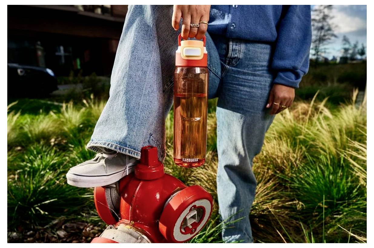 Egy személy egy nagy vízpalackot tart, amely "ZAMZAR" felirattal van ellátva, miközben az egyik lábát egy piros tűzcsapra helyezi egy füves területen.