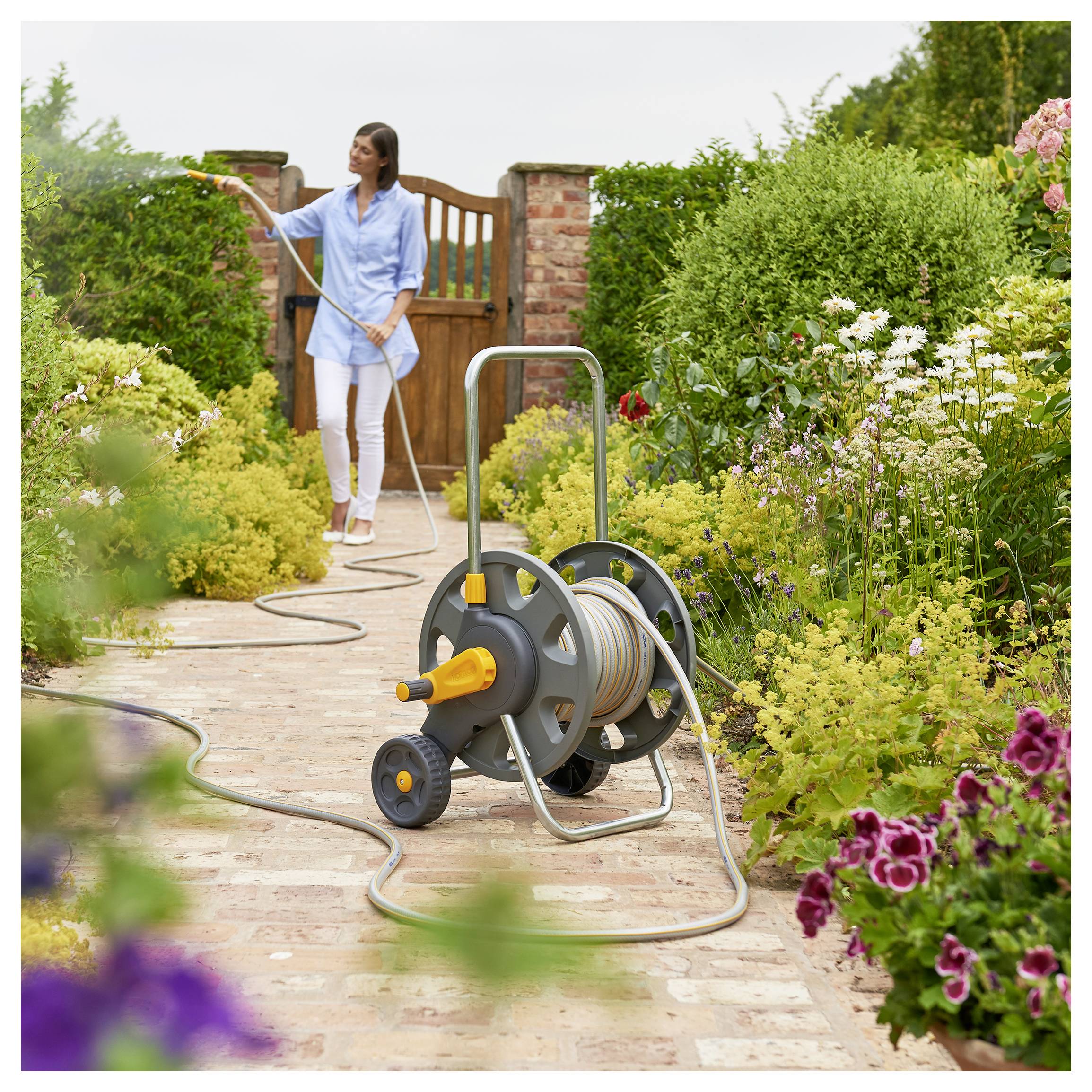 Egy személy locsolócsoron keresztül öntözi a kertbeli növényeket egy virágokkal és zöldellő növényzettel szegélyezett burkolat úton.