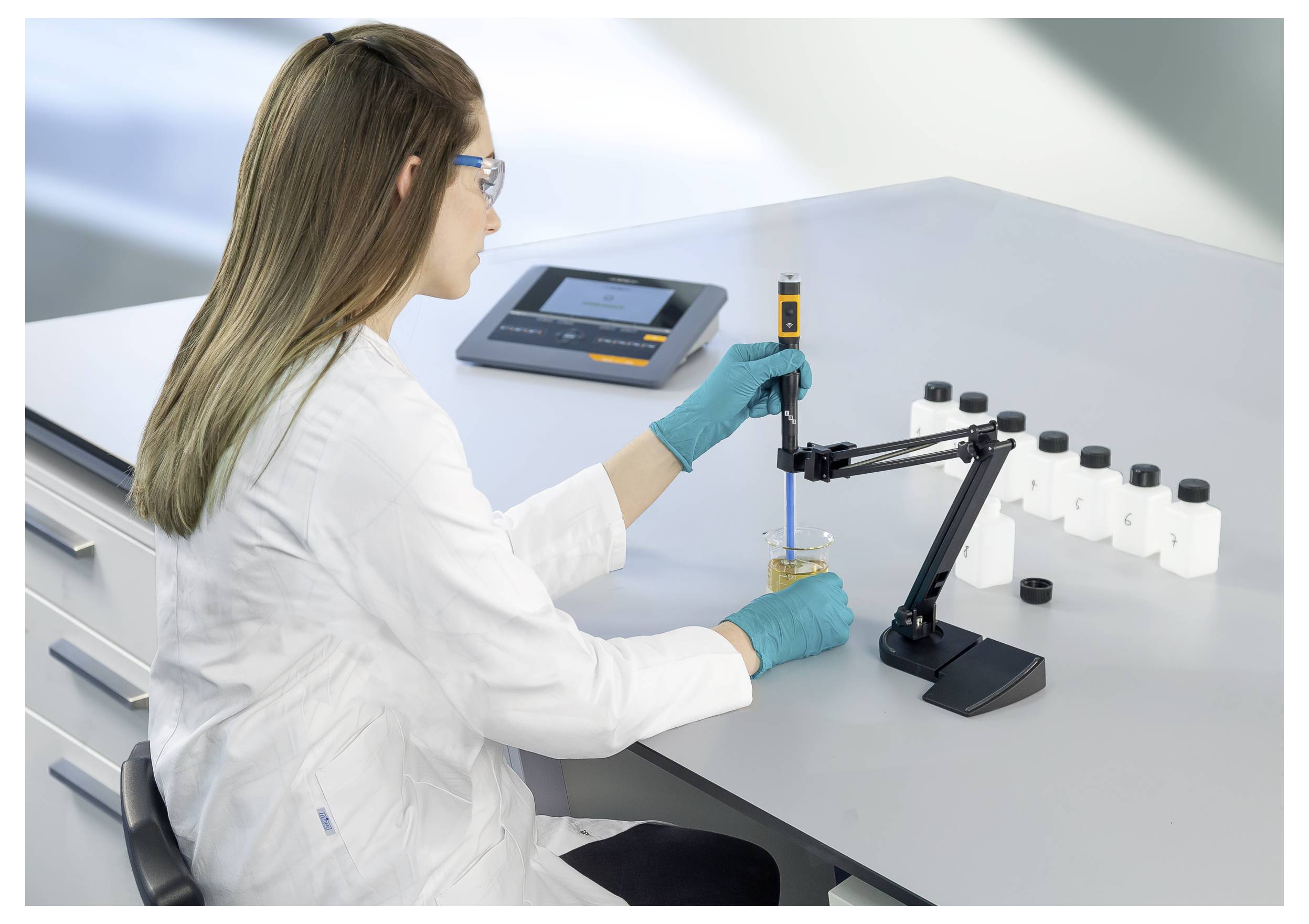 Egy fehér laborköpenyben lévő tudós egy digitális pipettát használ egy mintán. Az asztalon üvegek és egy készülék vannak, a háttérben laboratóriumi felszerelések láthatók.