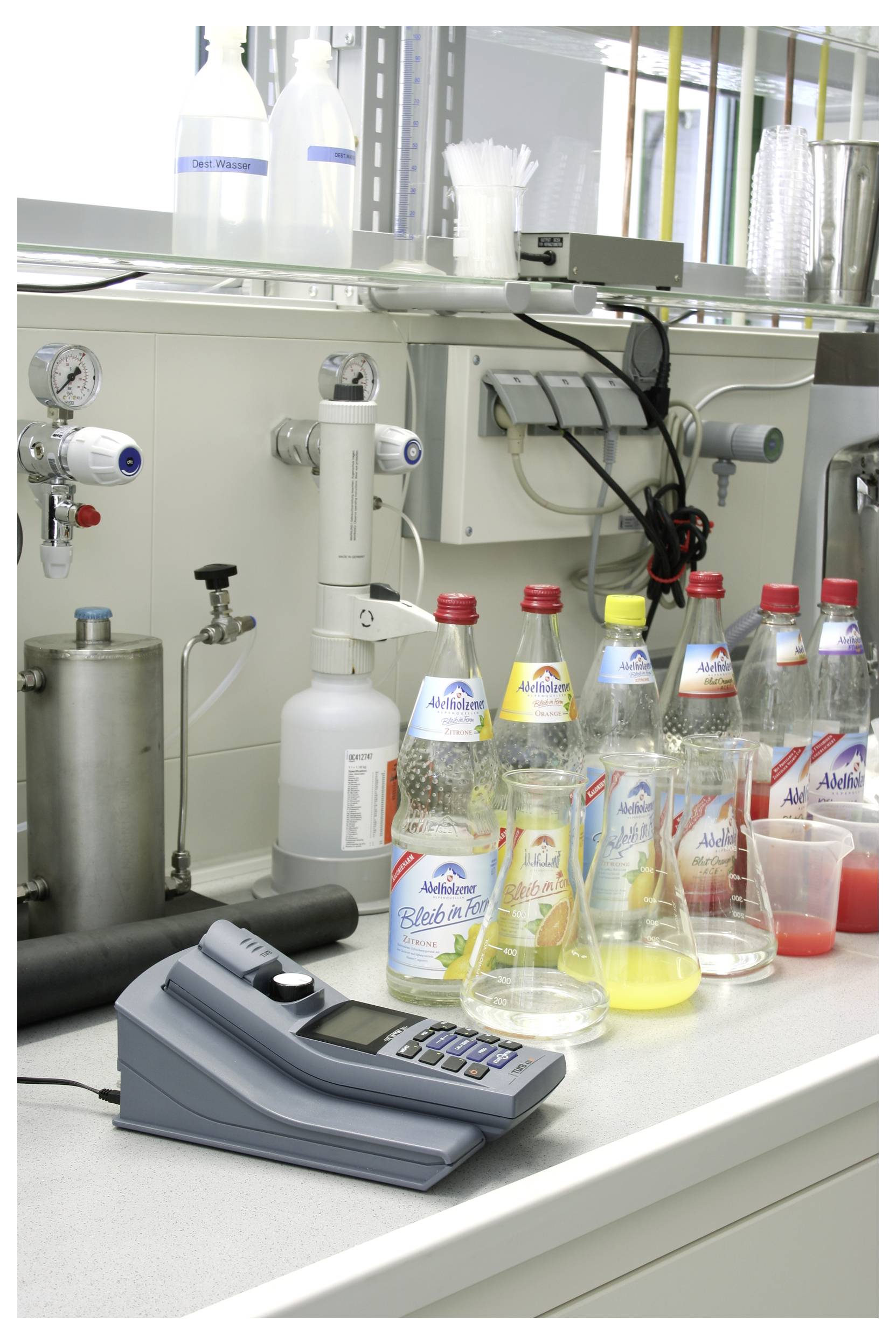 Laborátóriumi munkapult különböző Adelholzener ásványvízüvegekkel, mérőeszközzel és laboratóriumi berendezéssel, amely tesztelésre vagy elemzésre utal.