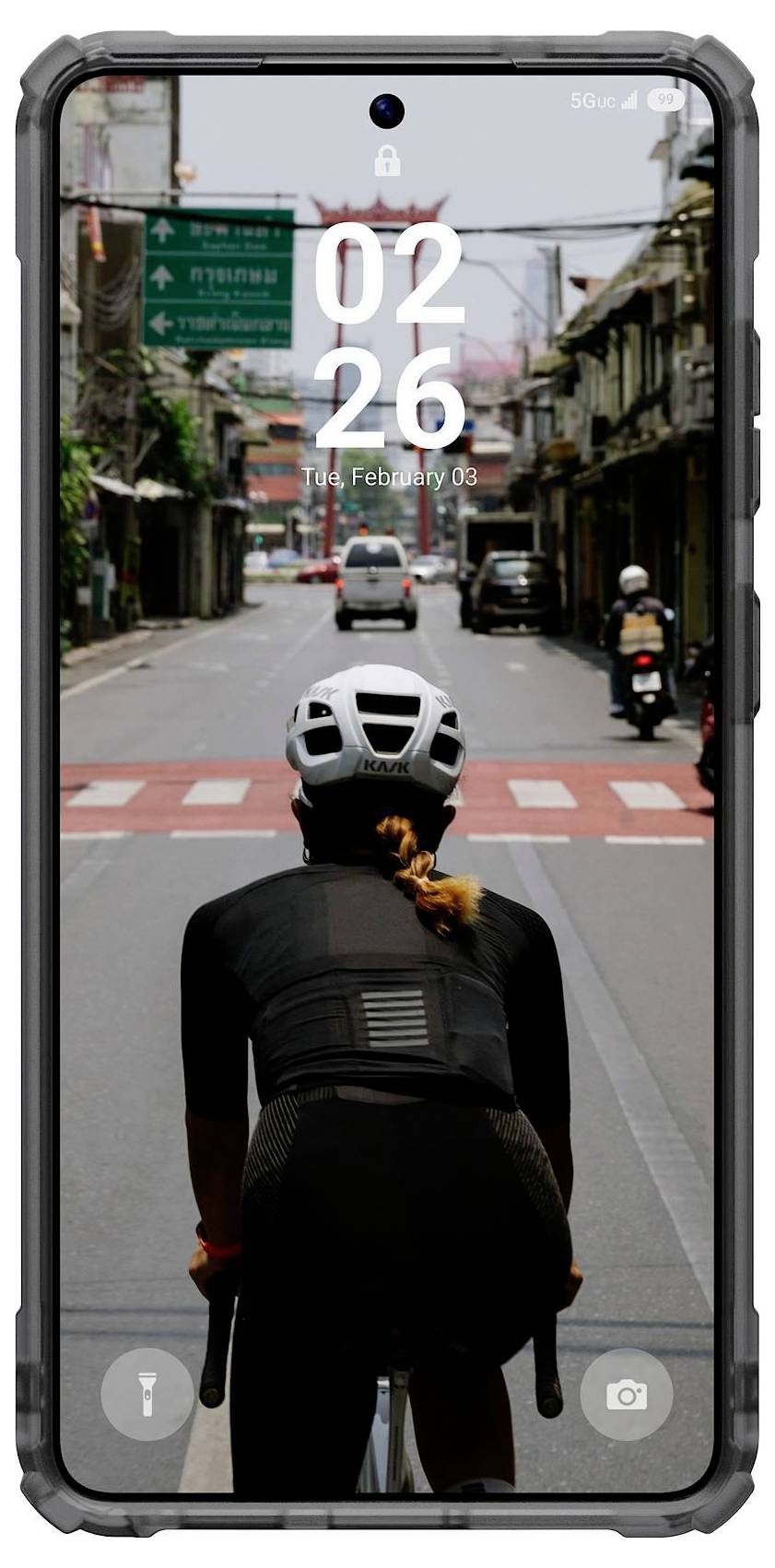 Kerékpáros egy kerékpáron lovagol egy városi utcán, amelyet épületek szegélyeznek, egy okostelefon képernyőjén látható, amely az időt 02:26-ként mutatja.