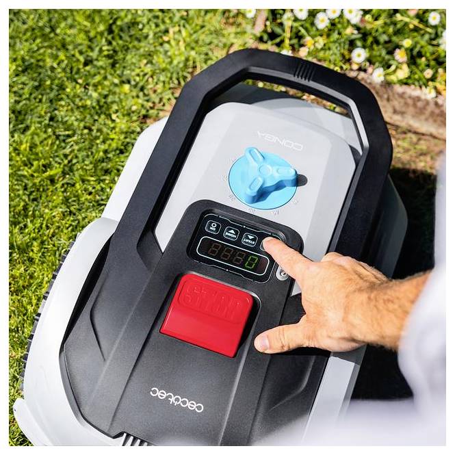 Egy személy keze megnyom egy gombot a robotfűnyírógép vezérlőpanelján, amely a füvön található, a háttérben virágok láthatók.