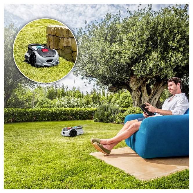 Egy személy egy kék kültéri kanapén relaxál, egy tabletet használ, miközben egy robotikus fűnyíró autonóm módon nyírja a füvet egy napos kertben.