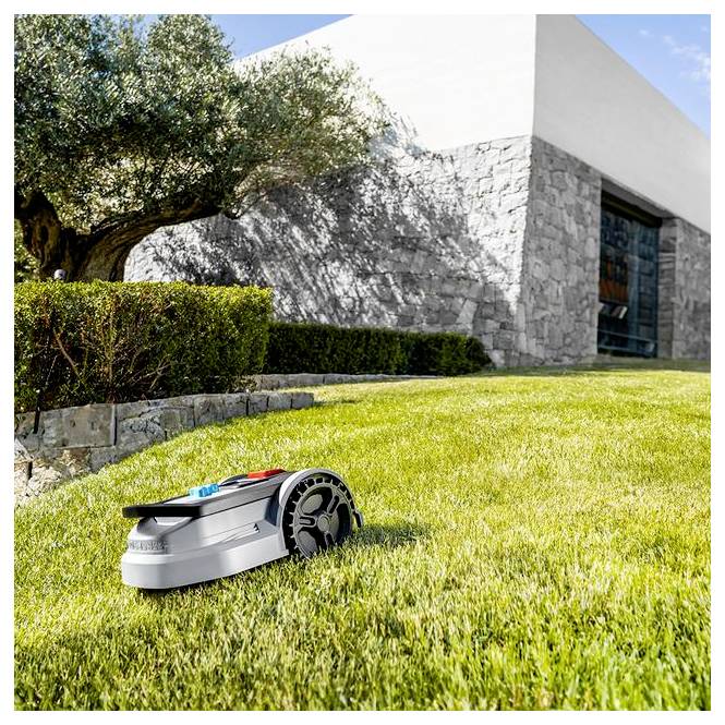 Egy robotfűnyírógép frissen kaszált füvön, egy kőfal és egy fa közelében, tiszta kék ég alatt, az automatizált gyepápolást szemléltető.