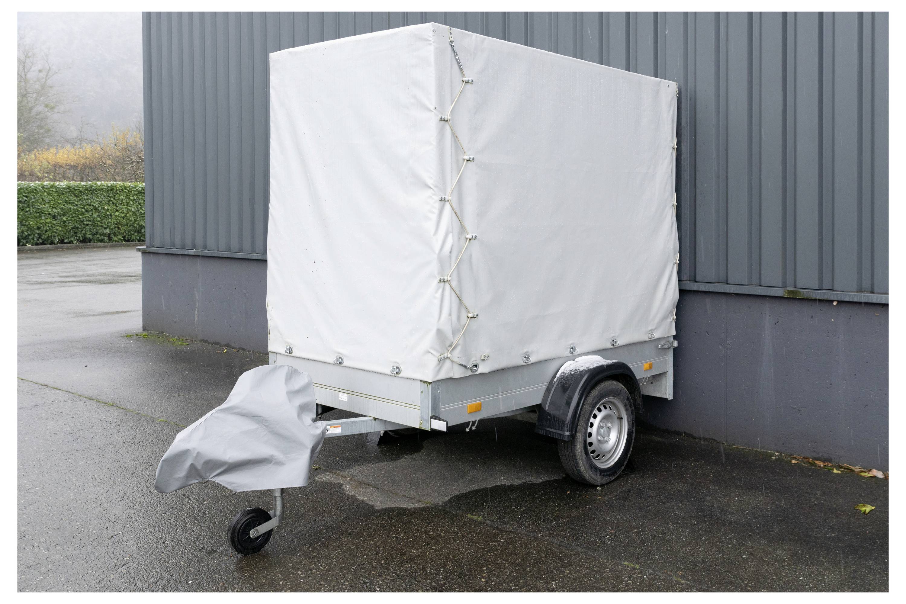 Egy kicsi, zárt utánfutó szürke ponyvatakarással a pakolyon egy sötétszürke épület mellett áll a nedves járdán.