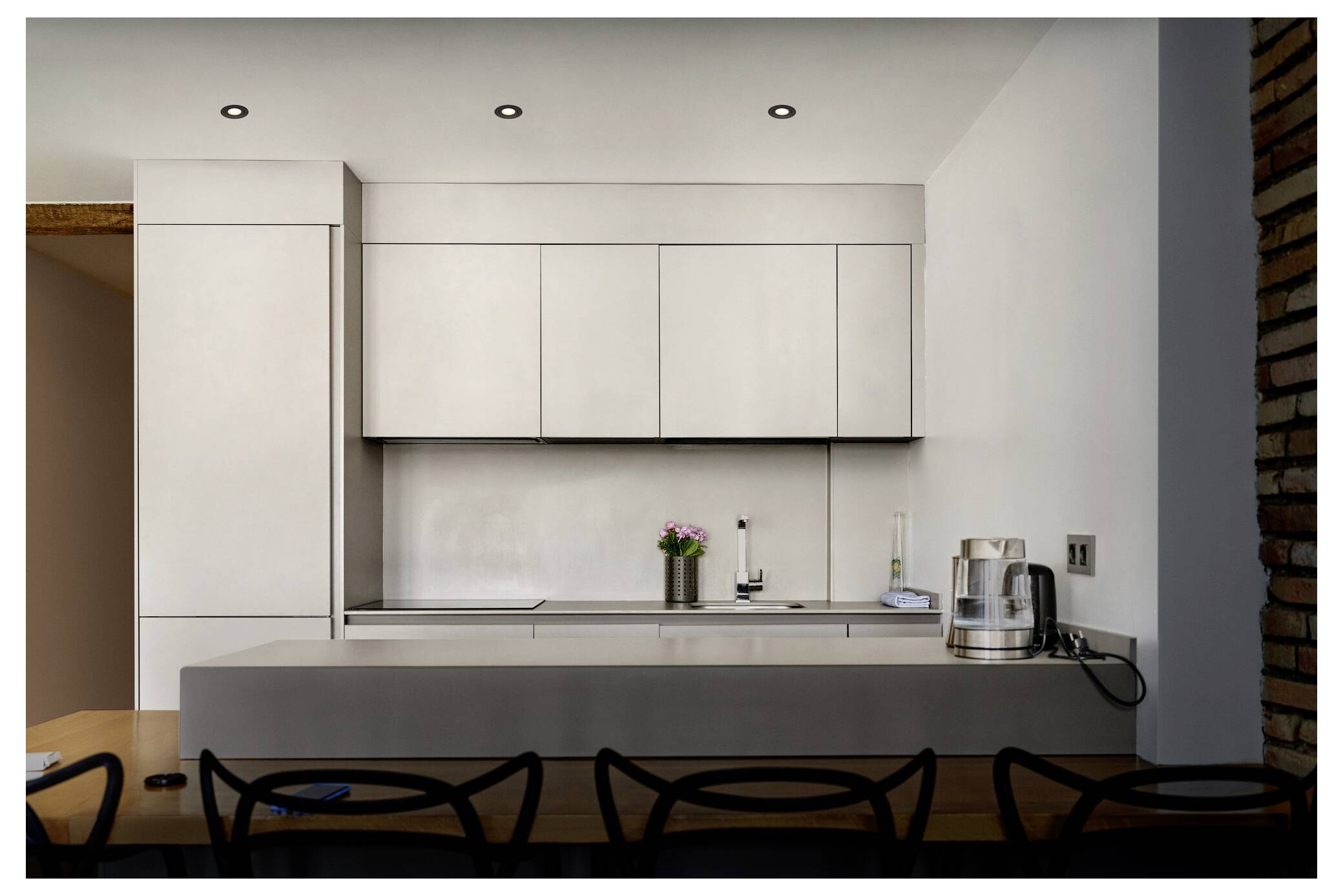 Minimalista modern konyha fényes fehér szekrényekkel, szürke munkalappal és beépített készülékekkel. A munkalap tetején egy kicsi növény és a konyha alapvető eszközei helyezkednek el.