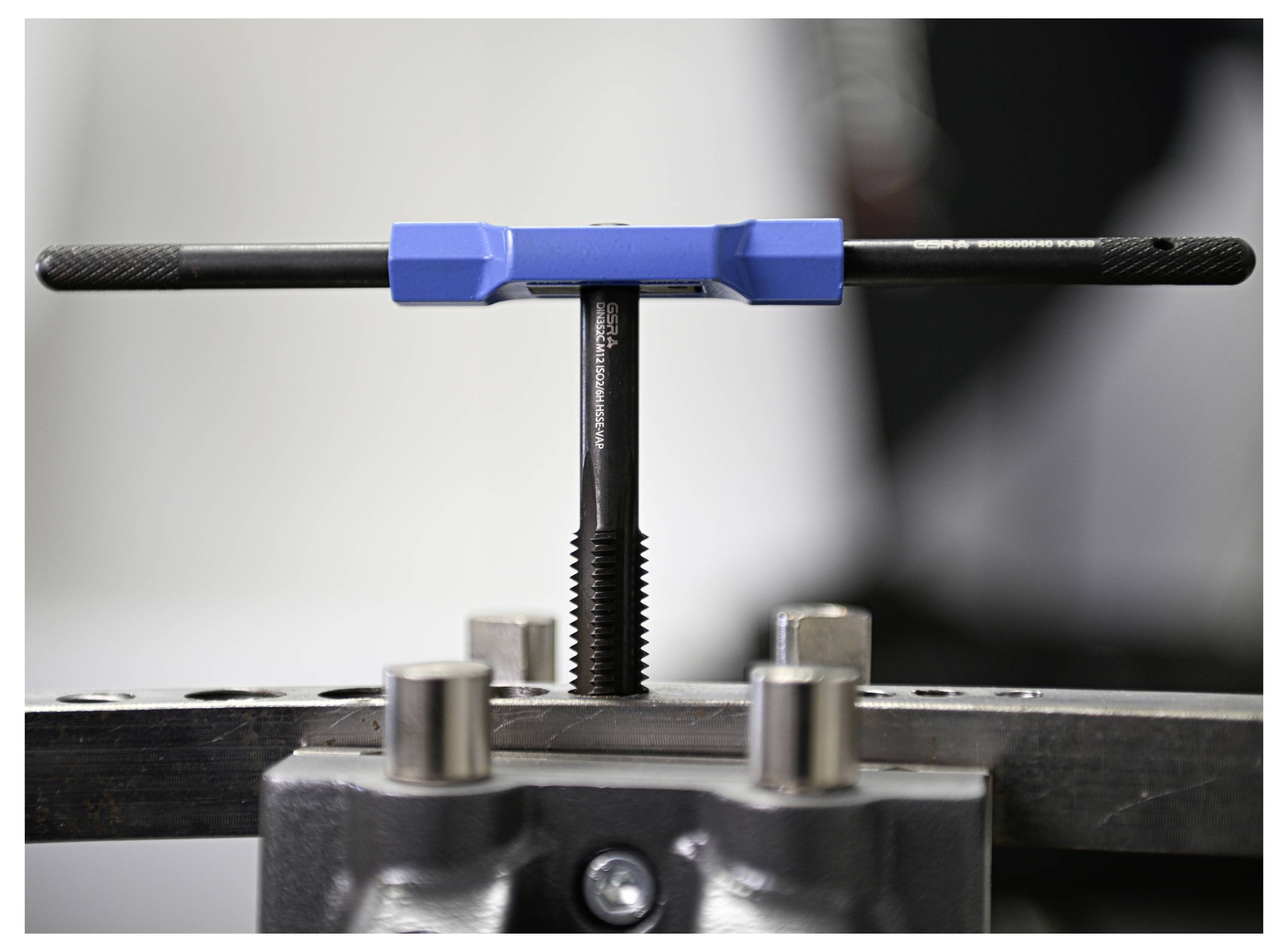Egy kék fogantyúval rendelkező csapmaró vízszintes helyzetben biztonságosan rögzítve van egy satu szerszámban, amely a menetezési vagy karbantartási munka beállítására utal.