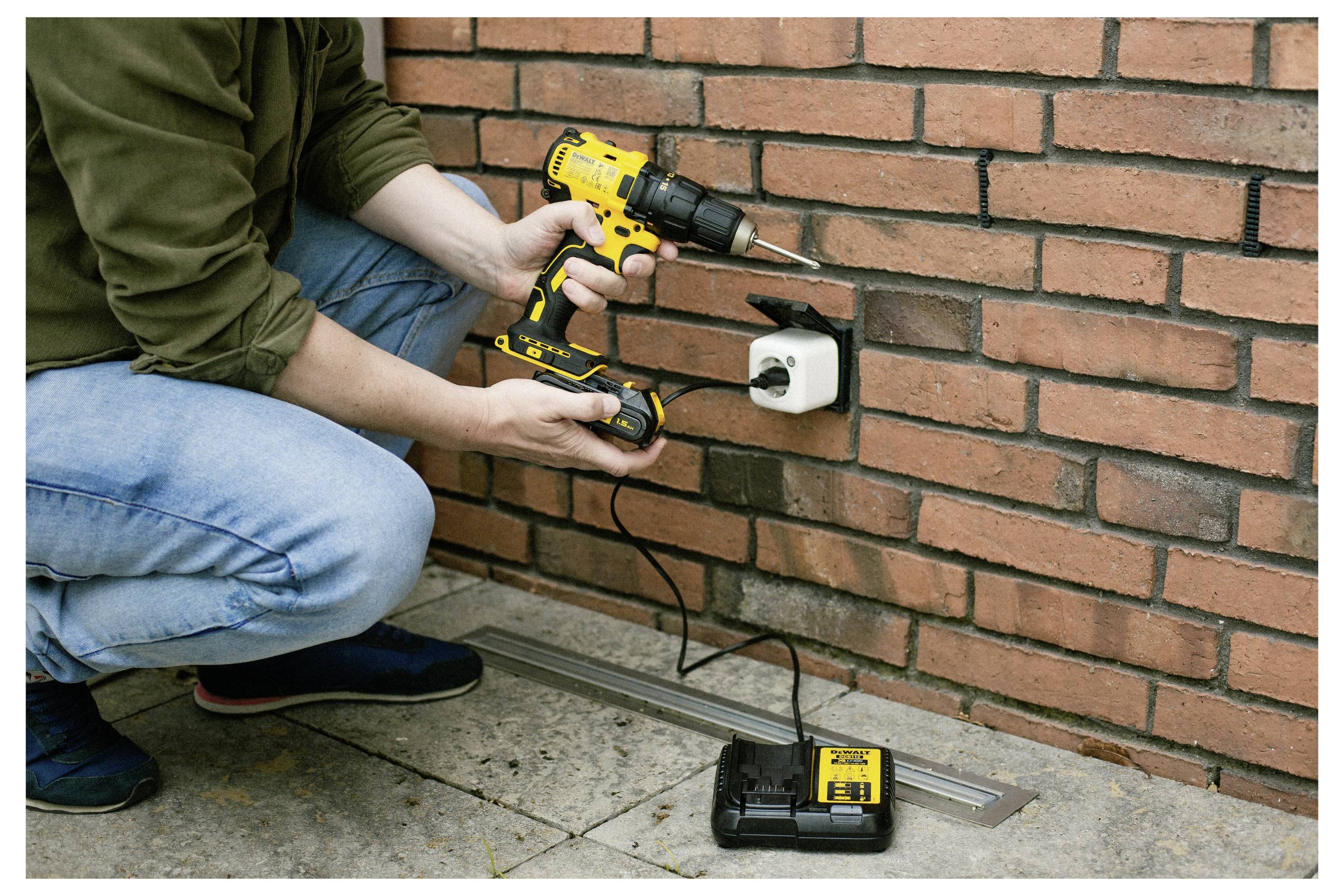 Egy személy egy sárga akkumulátoros fúrógépet használ egy téglafalon rögzítve egy eszközt, amely a DIY-lakásfelújítást mutatja. Az akkumulátor és a töltő a talajon látható.