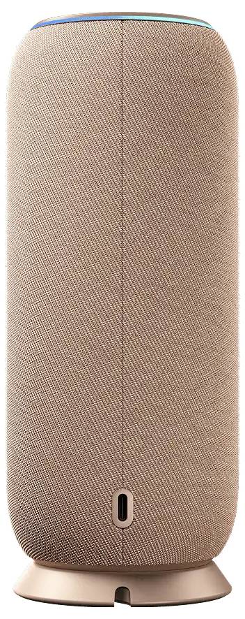 Magas hengeres textil hangszóró bézs színben, elölnézetből egy kerek talpzaton, kis jelzőfénnyel a közelben az alján.