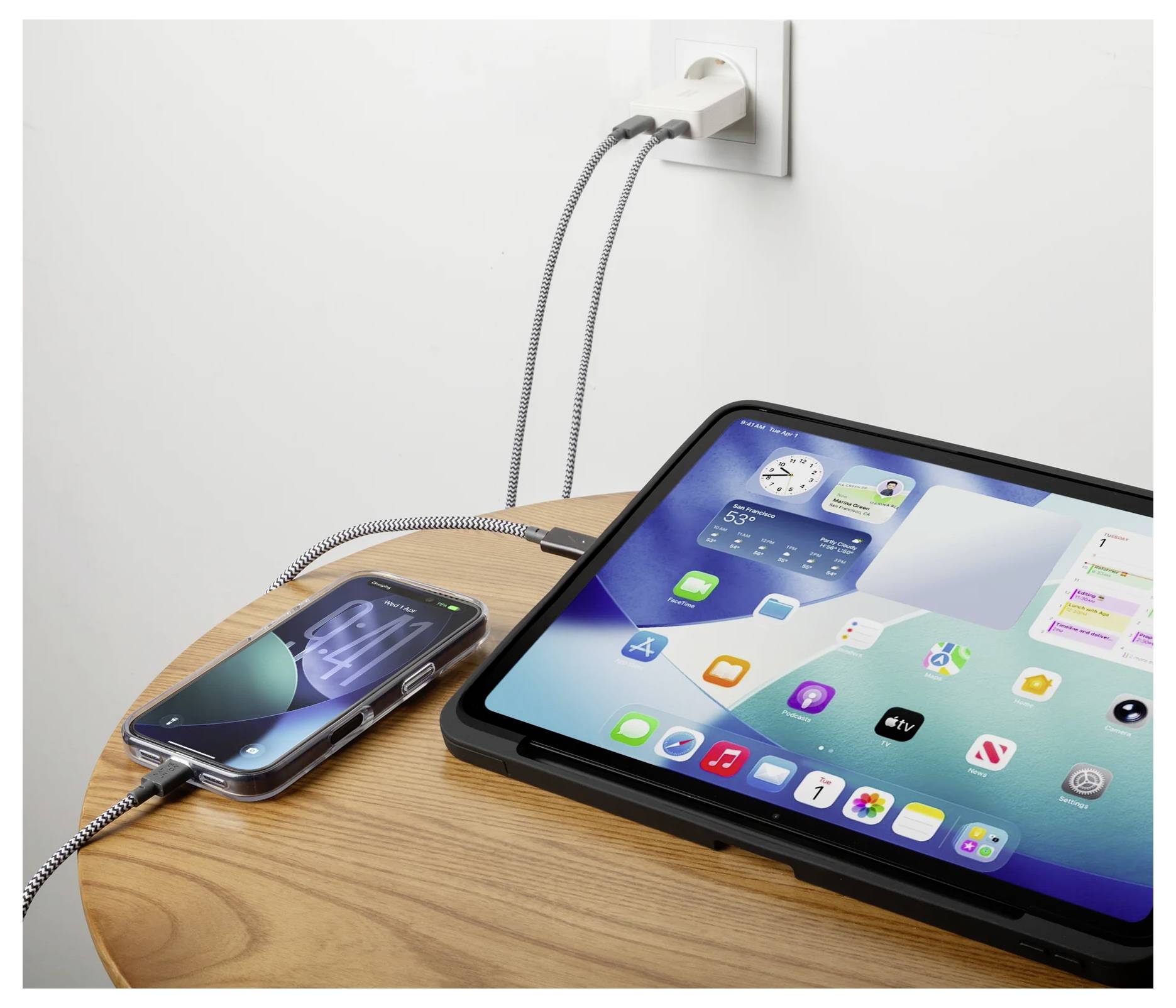 iPad és iPhone egymás mellett töltőznek egy fából készült asztalon, egy fali csatlakozóba dugva, kábelekkel csatlakoztatva.