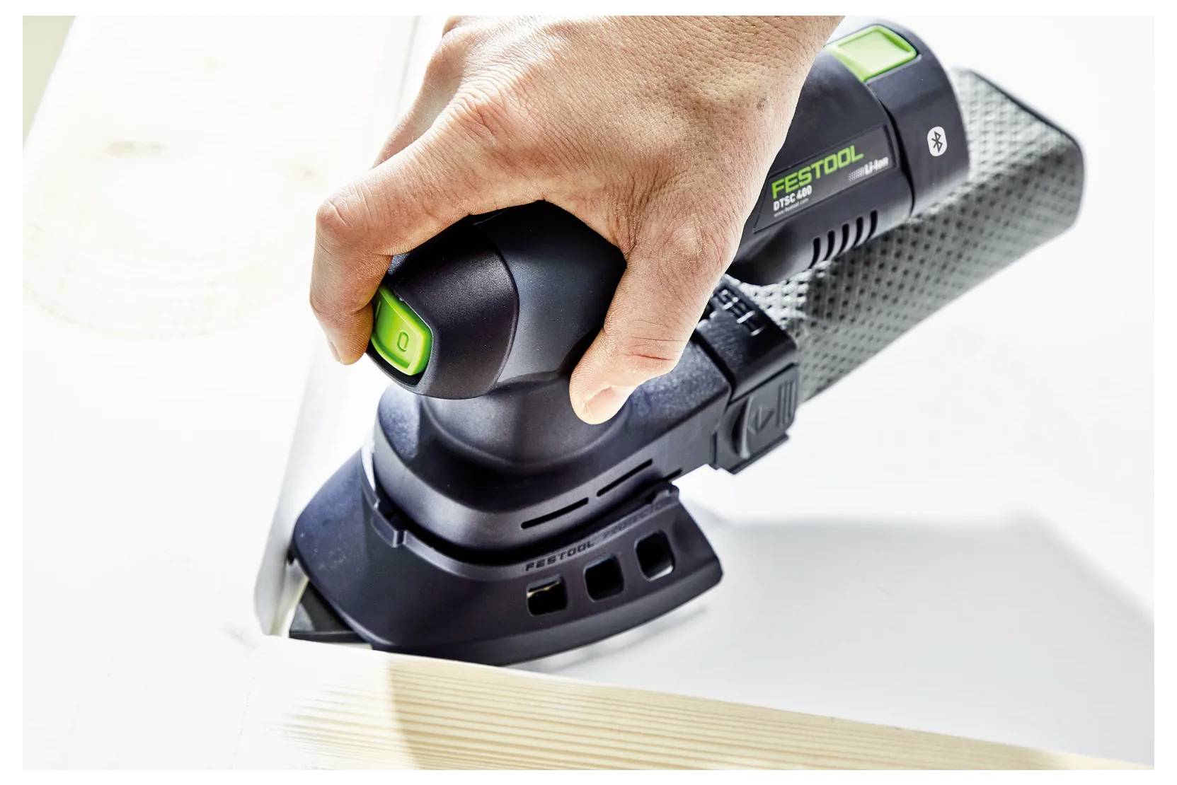 Kéz egy Festool elektromos csiszolóval egy fehér felületen, egy fa tábla szélét simítja.