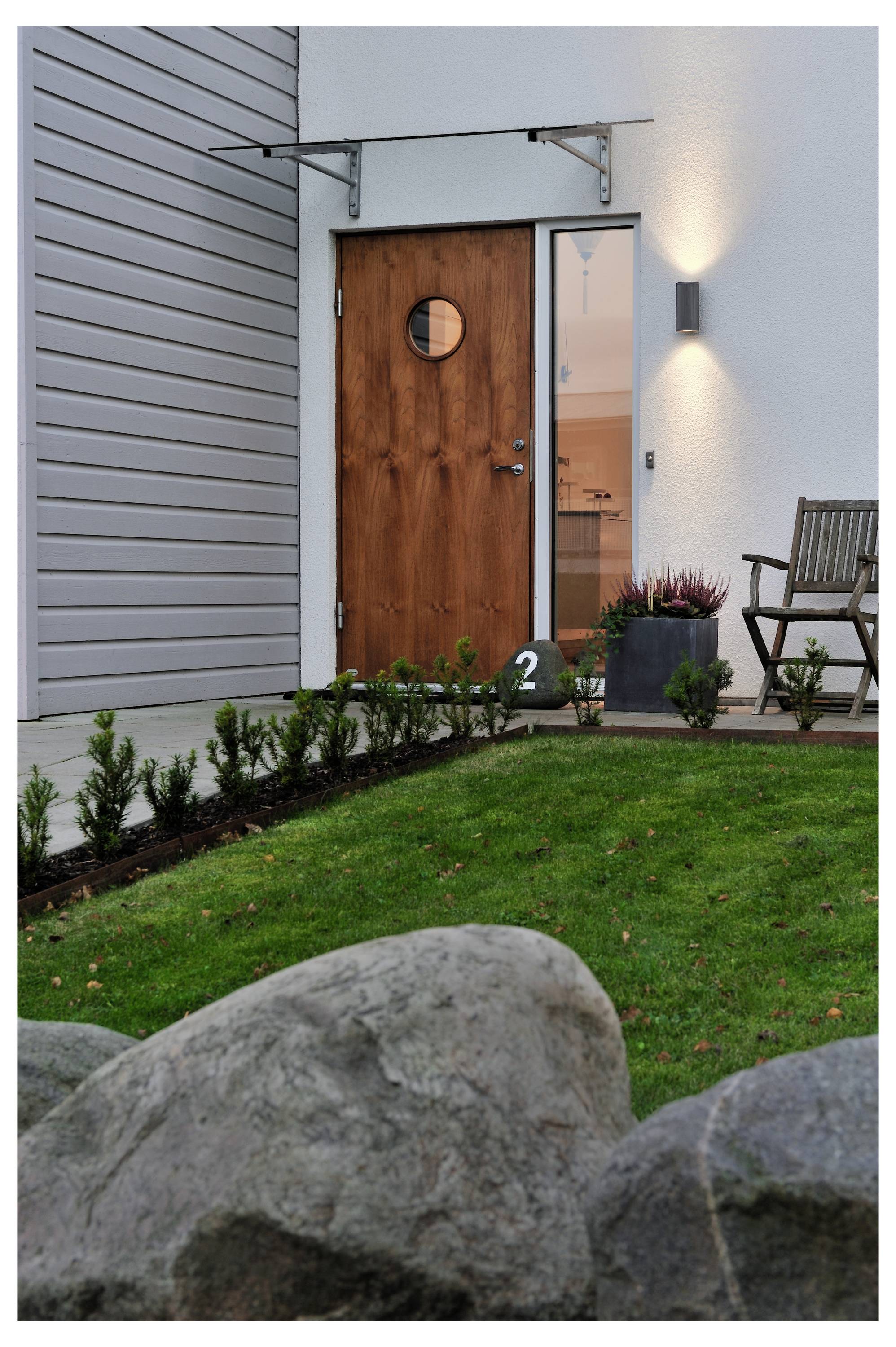 Modern házibejárat fából készült bejárati ajtóval, megvilágított falra szerelt fali lámpa, kis verandával, gyepvel, kövekkel és egy fotel az ajtó mellett.