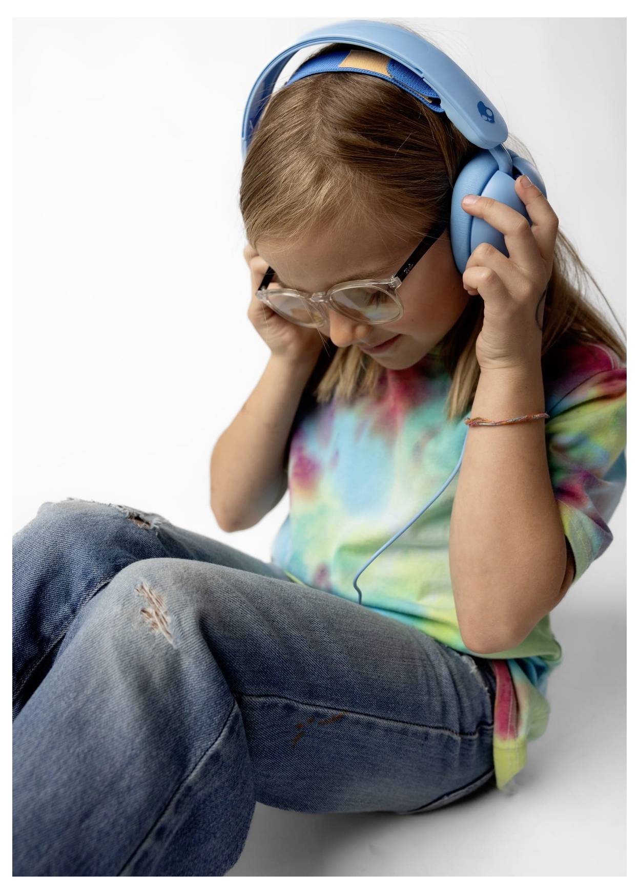Fiatal lány szemüveggel és kék fejhallgatóval keresztbe vetett lábakkal ül, lefelé néz, miközben beállítja azokat, pöttyös mintás pólót és farmert visel.