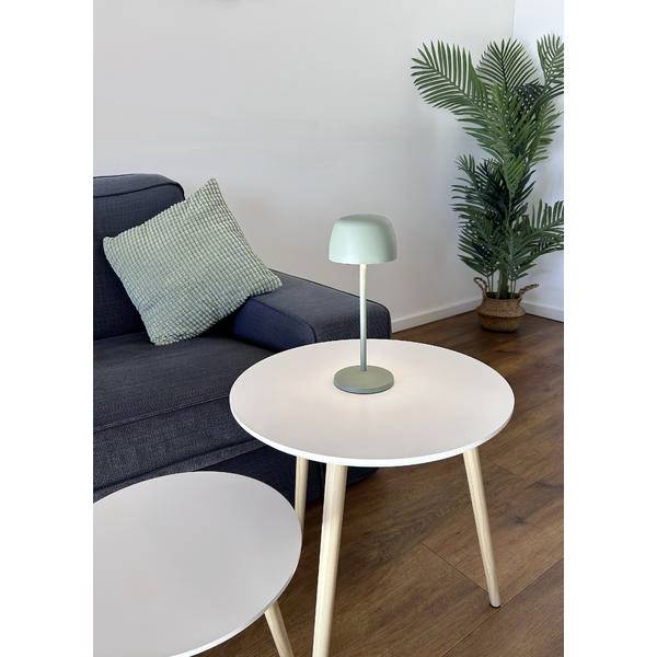 Modern nappali szoba sötét kanapéval, kerek fehér kávézóasztalokkal, egy kis asztalilámpa, és egy cserépbe ültetett pálma a fa padlózaton.