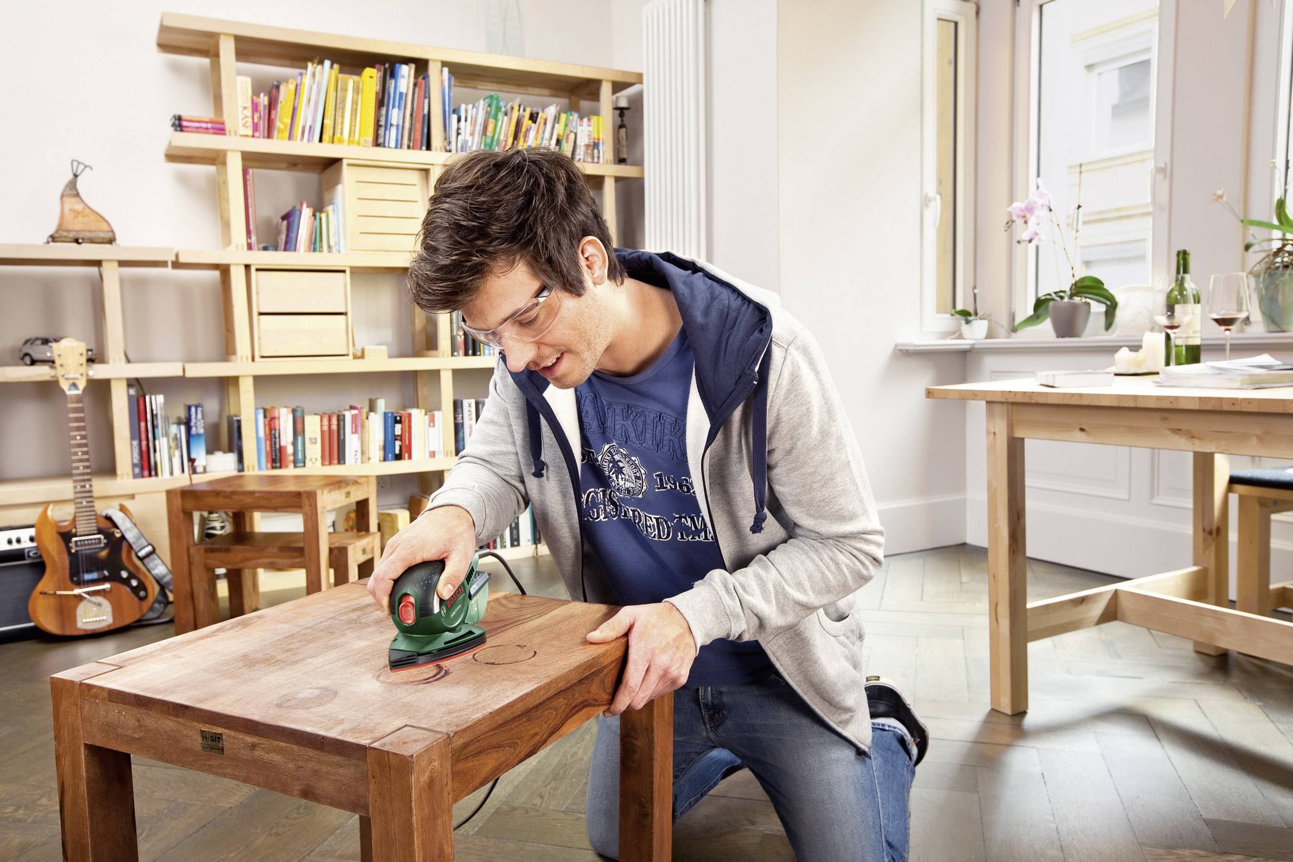 Egy férfi egy szerszámmal egy kisasztal fáját csiszolja egy világos szobában, amelynek háttérében polcok és egy asztal láthatók.