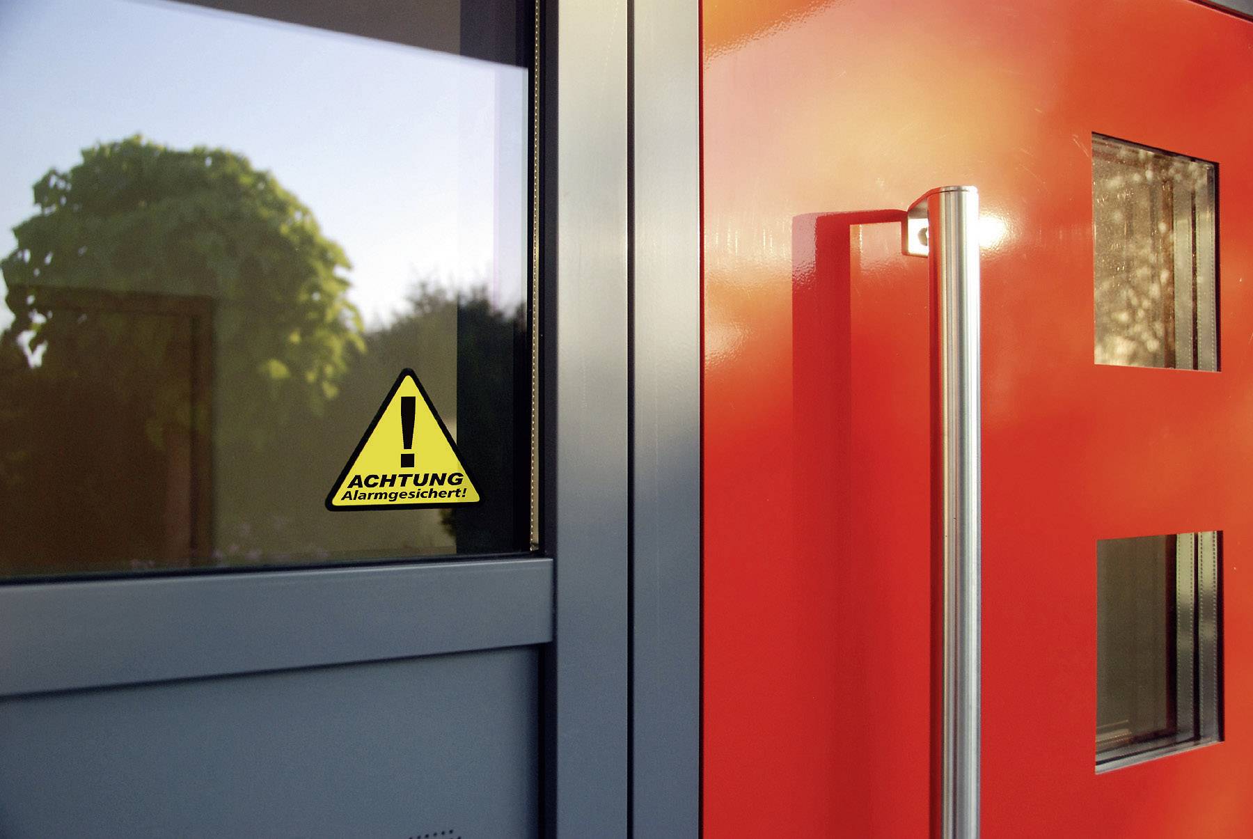 Egy piros ajtó üvegablakokkal és egy üvegablak mellette. Az üvegablakon egy sárga figyelmeztető tábla, rajta a felirat: 'Figyelem! Riasztóval védett'.