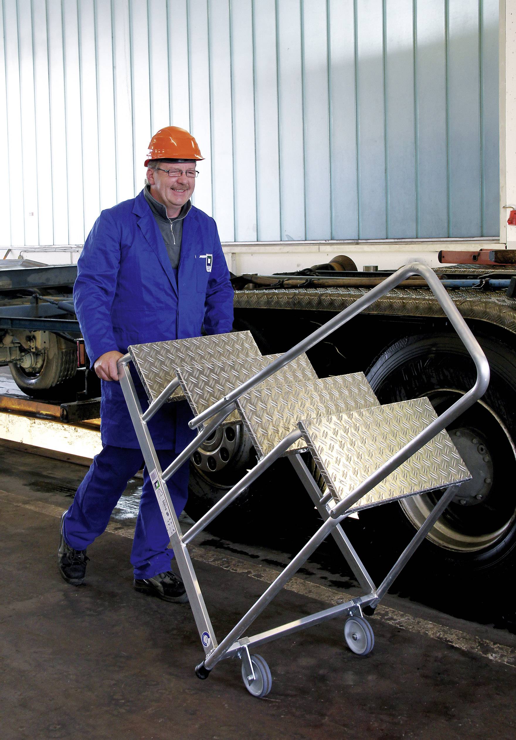 Egy munkásruhát és védősisakot viselő férfi egy mobil, kerekeken gördülő lépcsőt tol egy ipari környezetben.