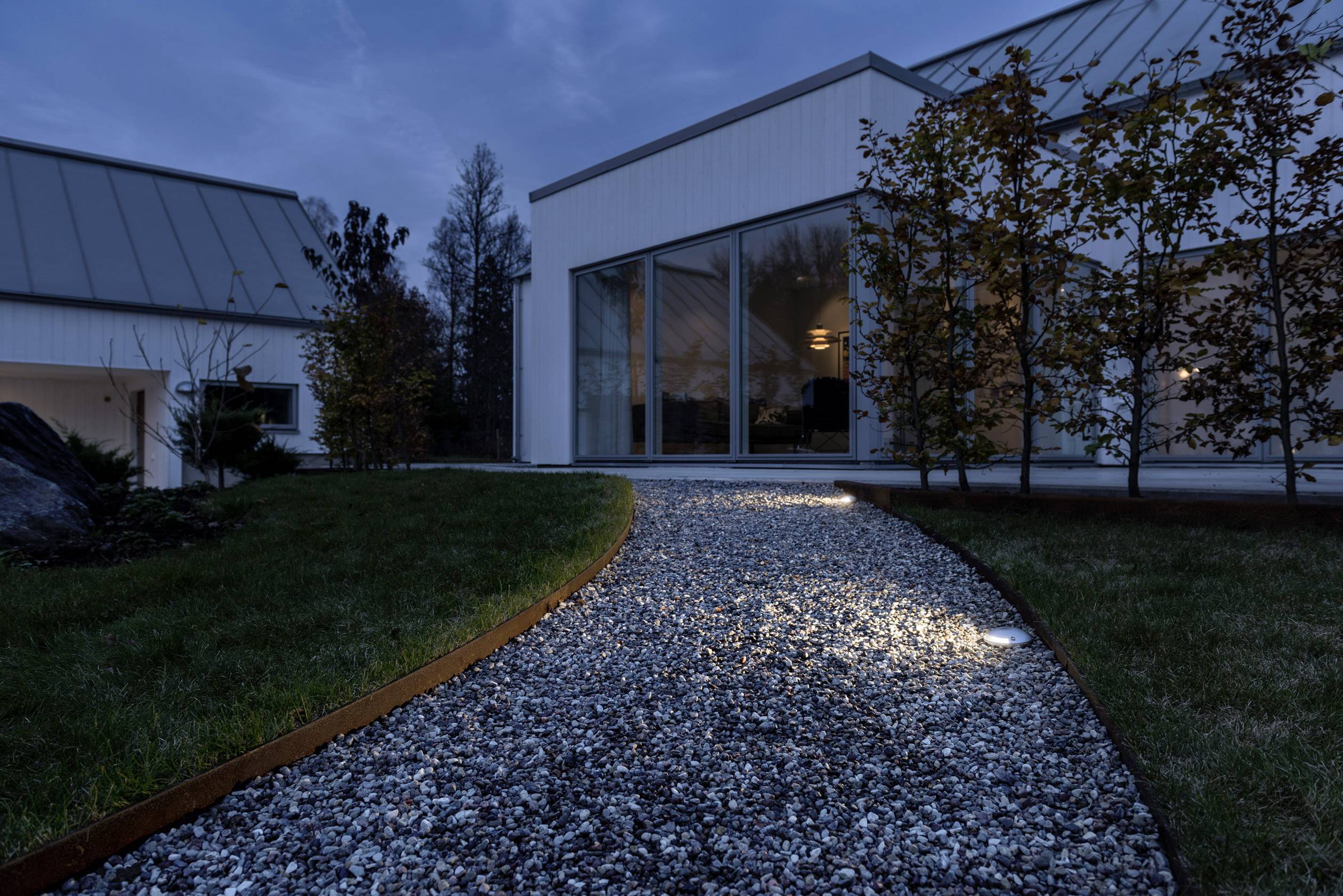 Egy kivilágított kavicsos ösvény vezet egy modern, üveg és beton házhoz, amely fák övezetében áll, alkonyatkor.