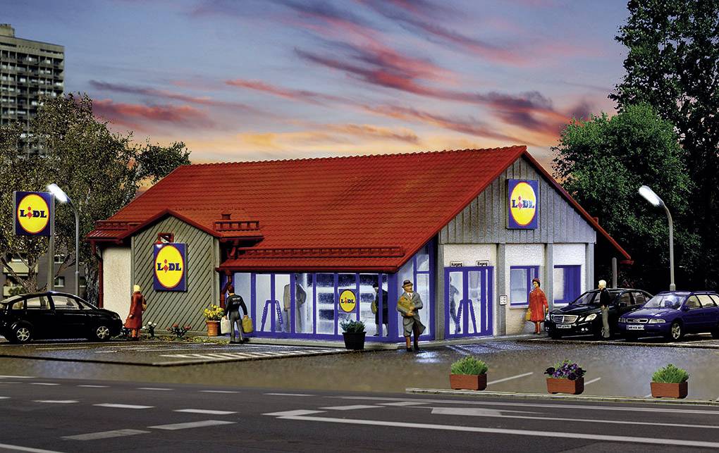 Egy kis, modern Lidl-üzlet piros tetővel naplemente idején. Emberek lépnek be az üzletbe, autók állnak a parkolóban.