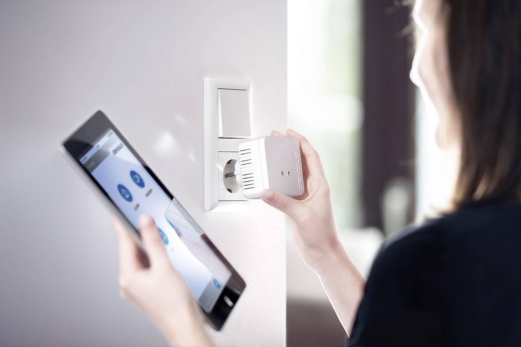 Egy személy egy intelligens dugaszolóaljzat-adaptert irányít egy táblagéppel; bemutatva a technika és az okos otthon közötti kapcsolatot.