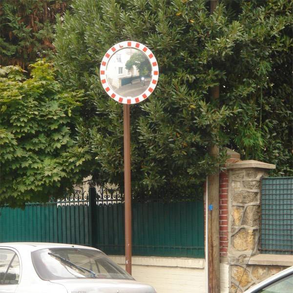 Egy útmenti közlekedési tükör visszatükrözi az út és a környező fák képét. Egy autó részben látható az előtérben.