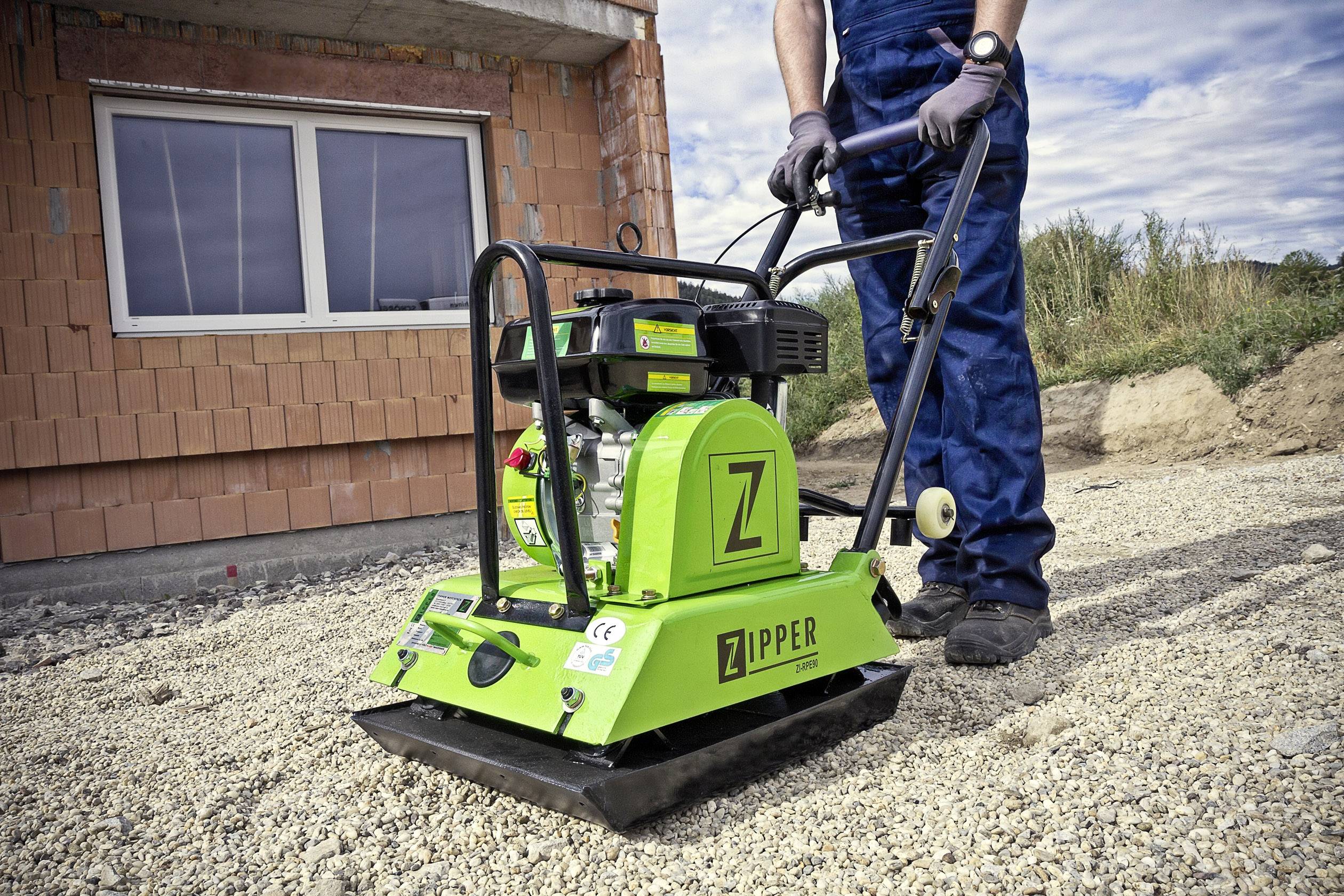 Egy munkásruhát viselő személy egy zöld lapvibrátort kezel egy kavicsos felületen egy nyers épület előtt.