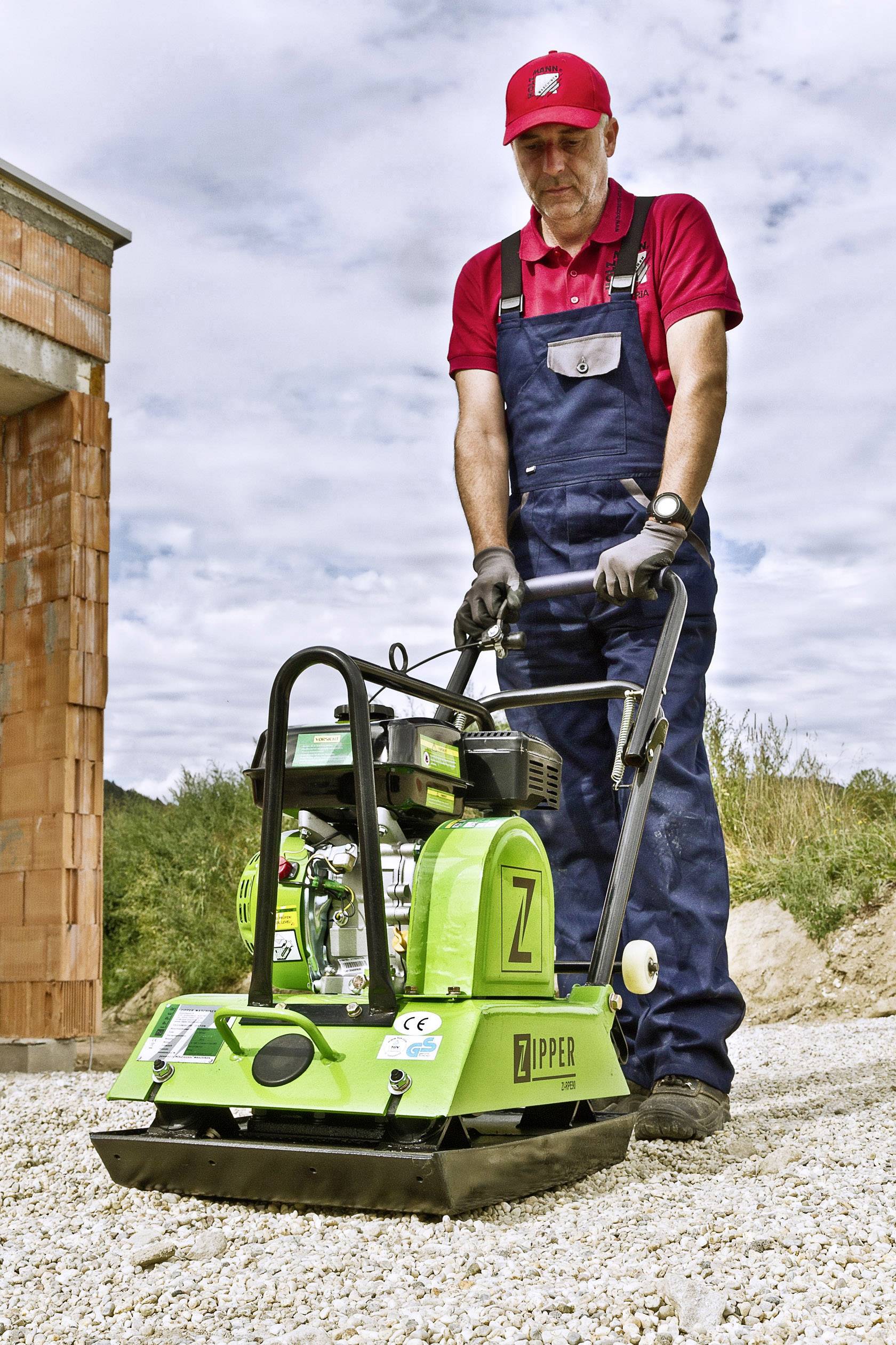 Egy munkás piros pólóban és kék overall-ban kezel egy zöld vibrációs lemezt egy kavicsos talajon egy épülő ház közelében.