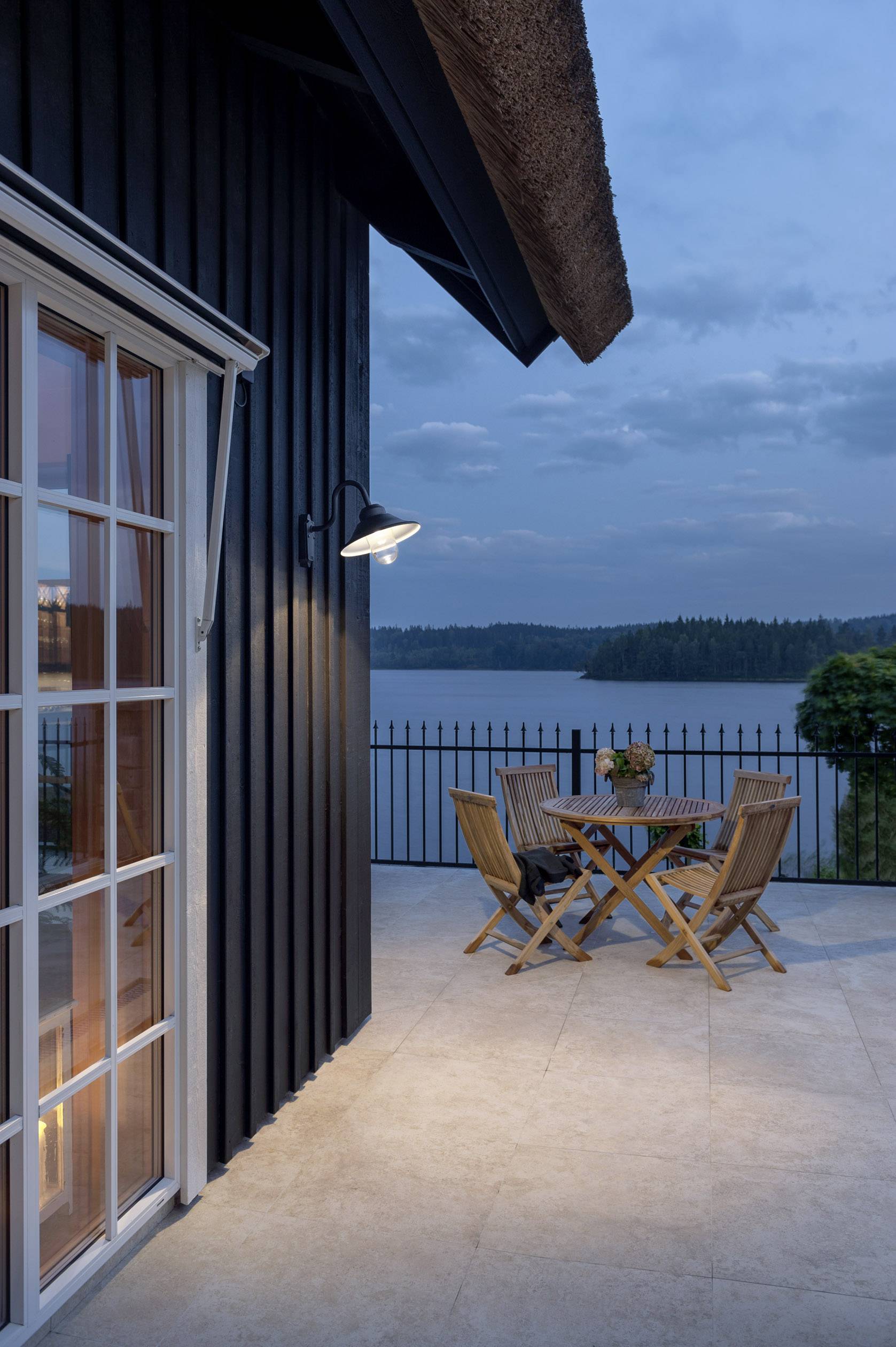 Terasz asztallal és székekkel, kilátással egy tóra alkonyatkor, egy házon lévő kültéri lámpa által megvilágítva.