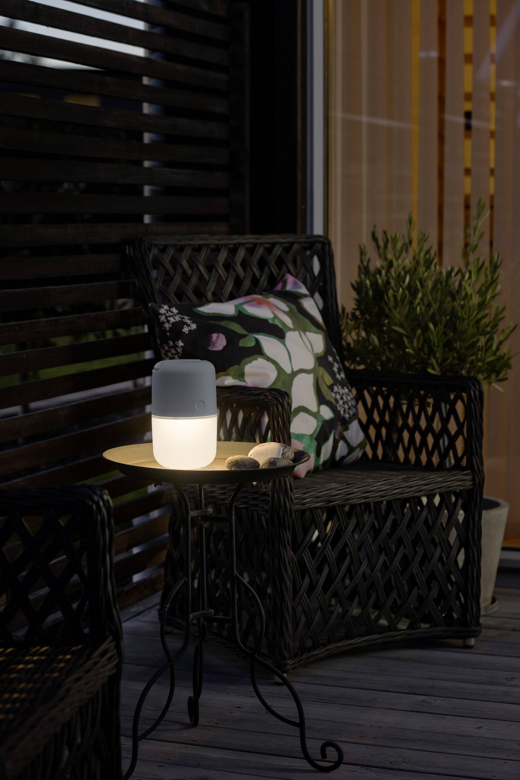 Egy megvilágított hordozható hangszóró áll egy asztalon egy virágmintás szék mellett egy teraszon alkonyatkor.