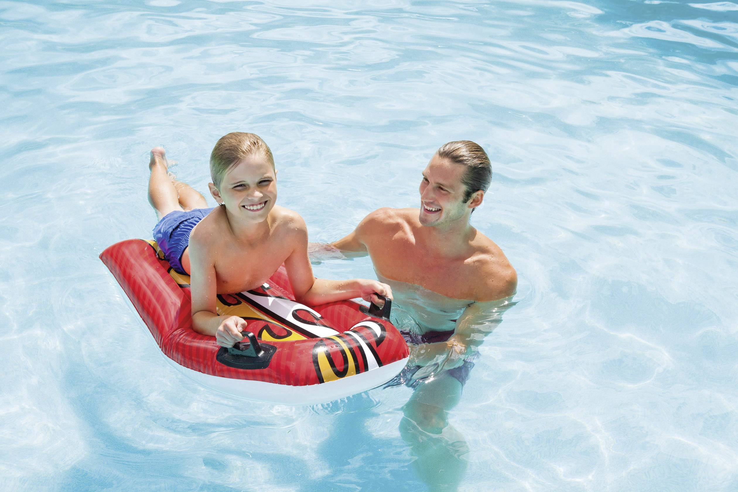 Egy mosolygó gyerek felfújható deszkán fekszik a medencében, egy felnőtt támogatja és játszik vele.